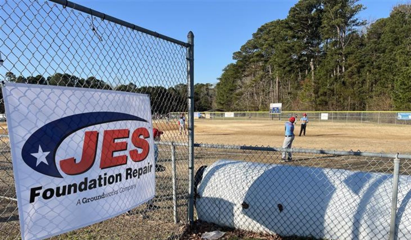 A baseball field with players and a "JES Foundation Repair" banner on a chain-link fence in the foreground.