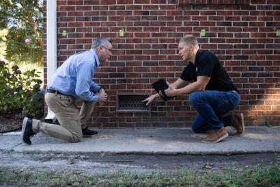 Two men kneel by a brick wall, examining a crawl space vent.