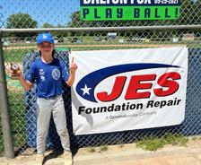 A young baseball player holds a trophy and points up, standing by a JES Foundation Repair banner at a baseball field.
