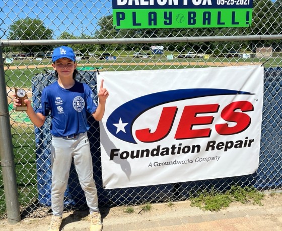A young baseball player holds a trophy and points up, standing by a JES Foundation Repair banner at a baseball field.