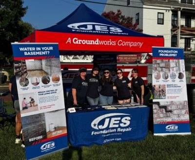 Five people stand at a JES Foundation Repair booth with banners and a red tent at an outdoor event.
