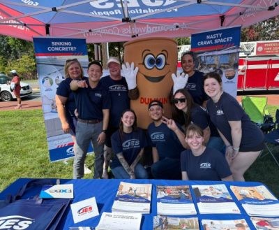 A group of people in JES shirts pose and smile with a Dunkin’ mascot at an outdoor event booth.