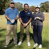 Three people in golf attire standing on a sunny golf course, smiling at the camera.