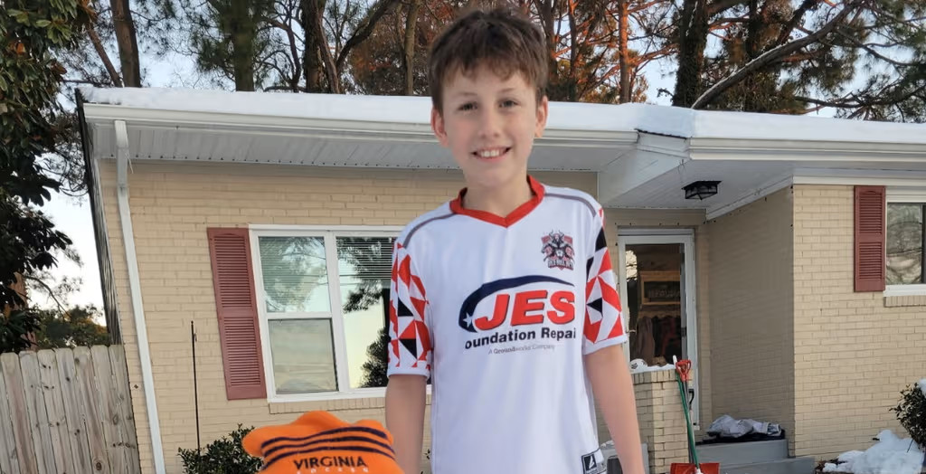 Smiling boy in a white soccer jersey stands outside a house with snow on the ground and roof.