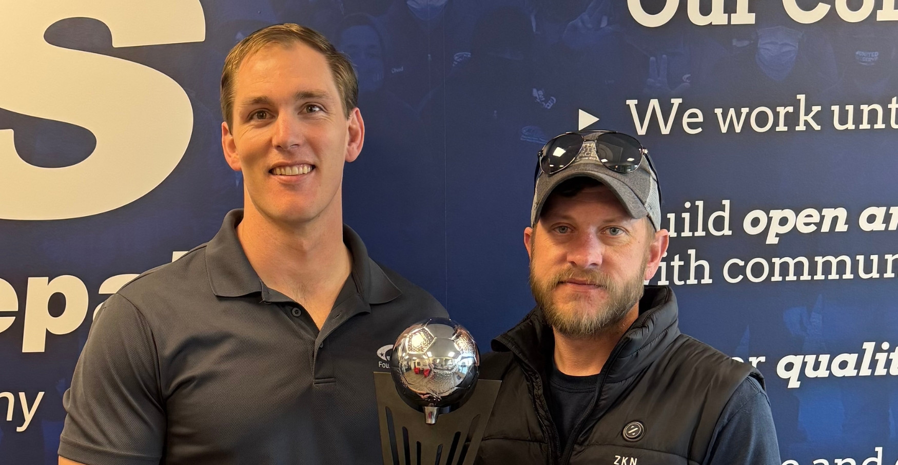 Two men standing together indoors, one holding a trophy with a silver soccer ball on top, smiling at the camera.