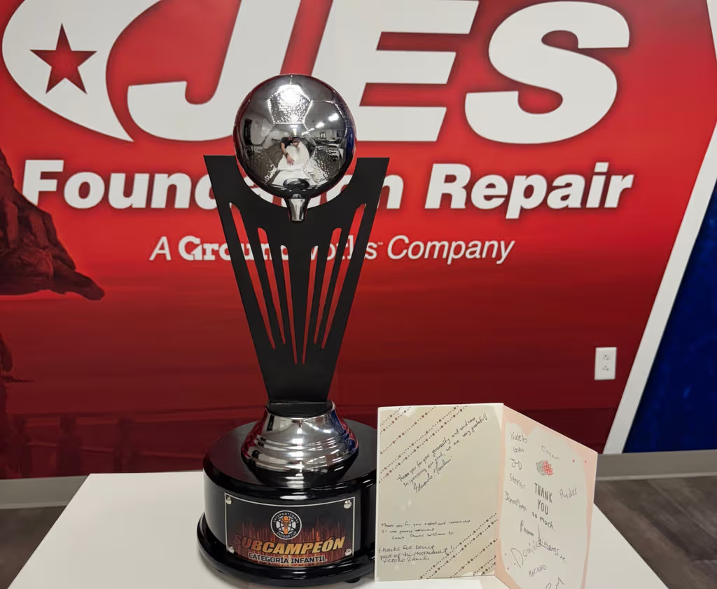 Silver soccer trophy and a greeting card on a table in front of a red JES Foundation Repair sign.