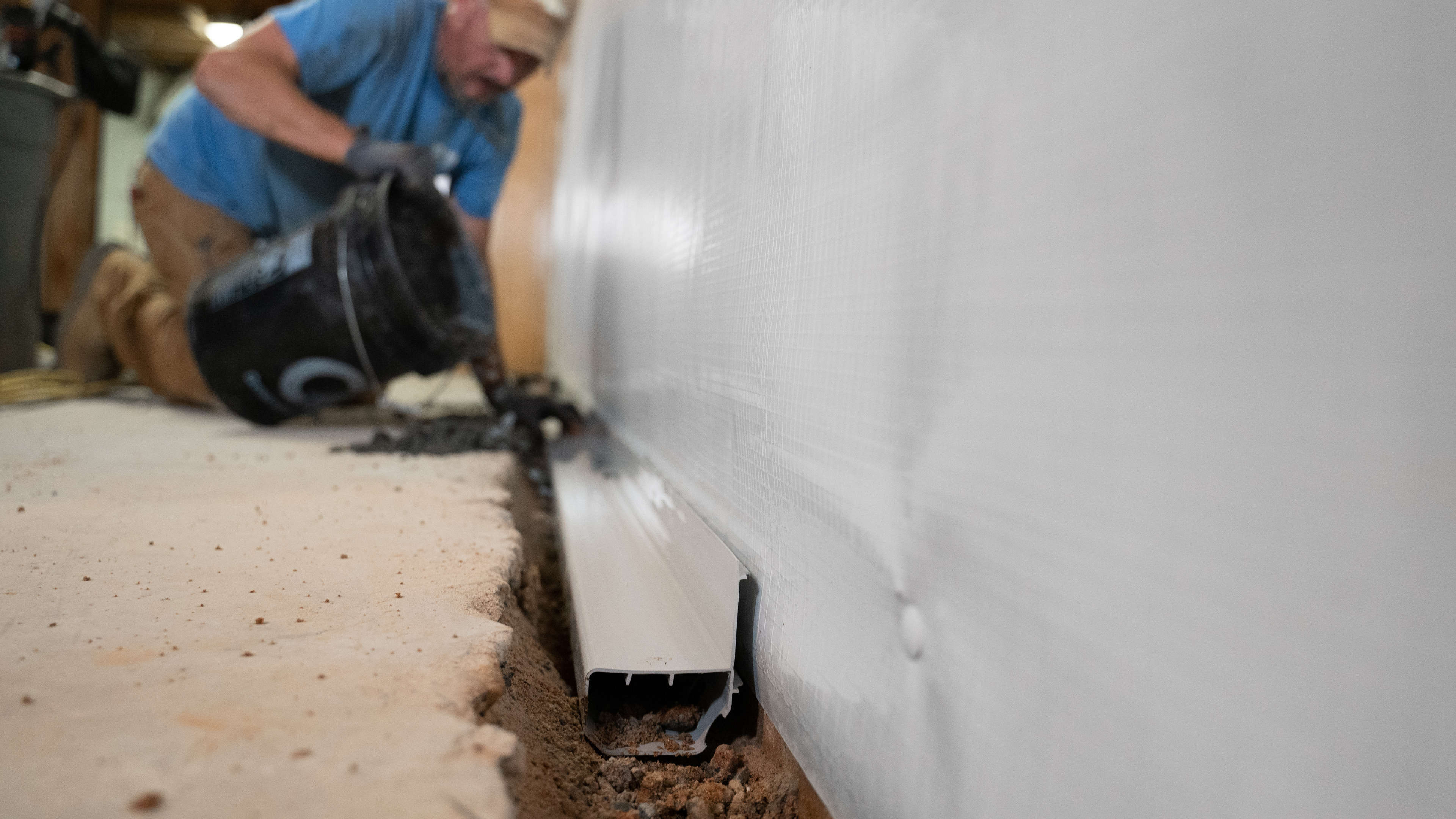 A worker pours gravel into a trench along a basement wall with a drainage channel installed.