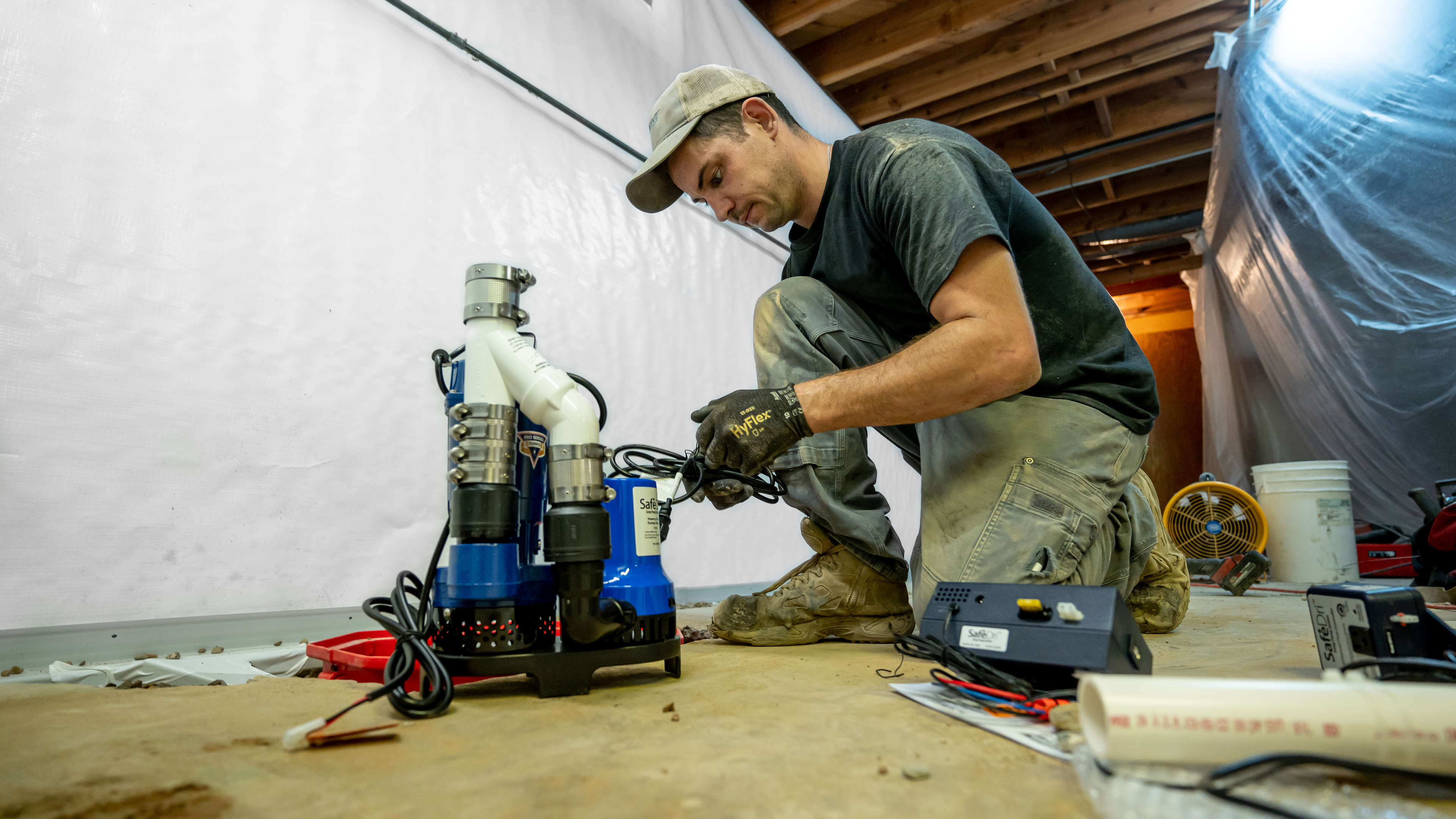A worker kneels on the floor, assembling or repairing a sump pump in a basement or crawl space.