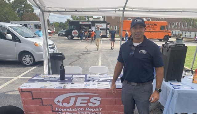 A man stands at a JES Foundation Repair booth at an outdoor event with food trucks in the background.