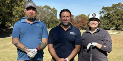 Three people in golf attire stand together on a golf course, smiling on a sunny day with trees in the background.