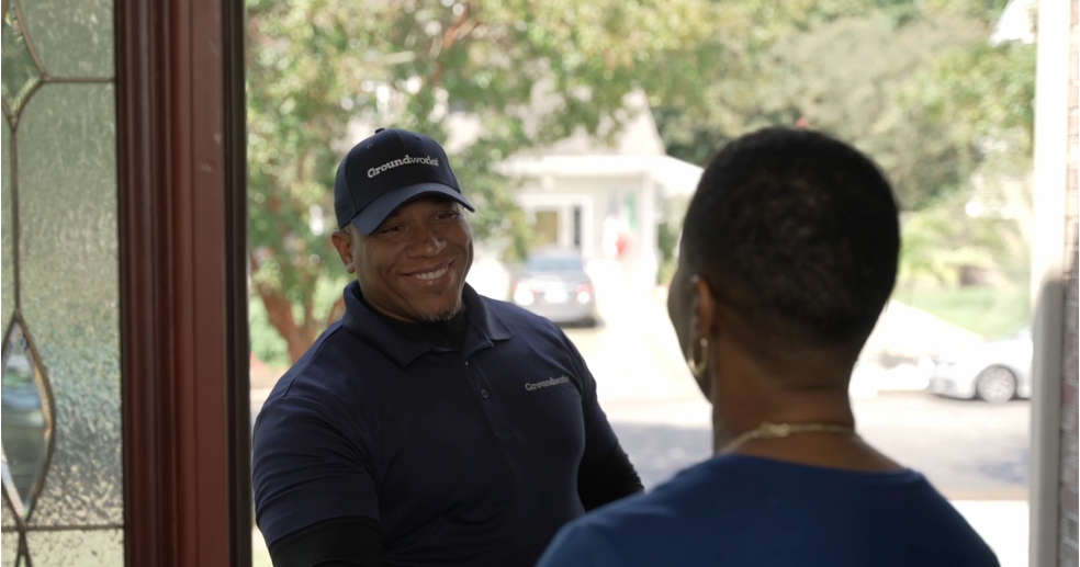 A smiling delivery person greets a woman at her front door on a sunny day.