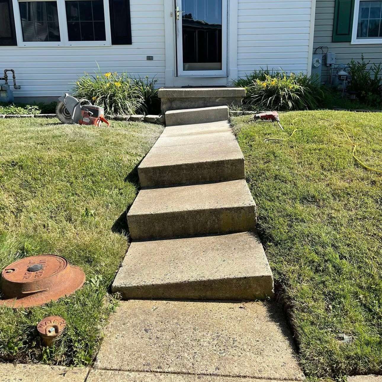 Concrete steps leading to a house entrance with grass on both sides and gardening tools nearby.