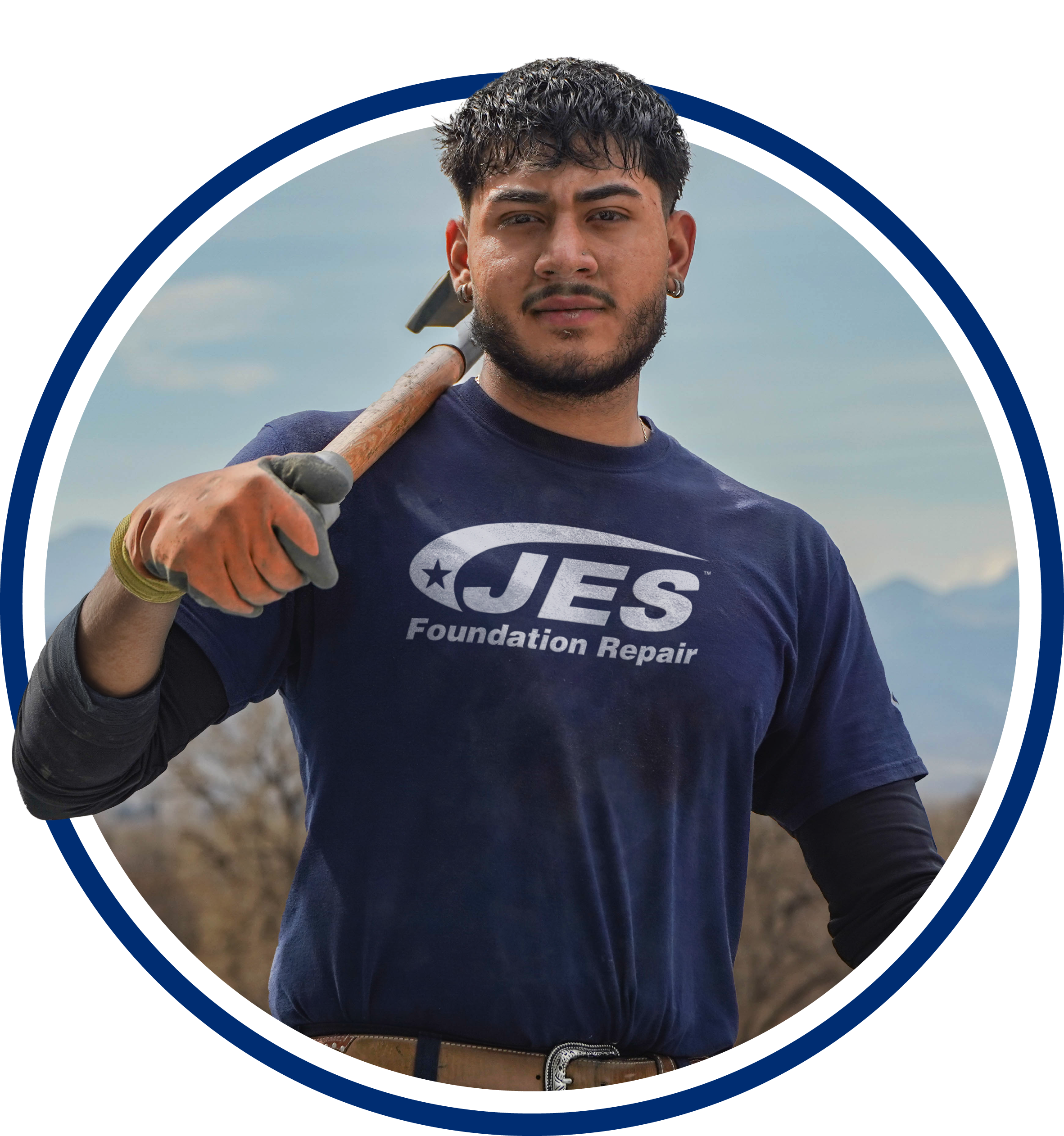 A man in a JES Foundation Repair shirt holds a tool over his shoulder, standing outdoors with mountains behind him.