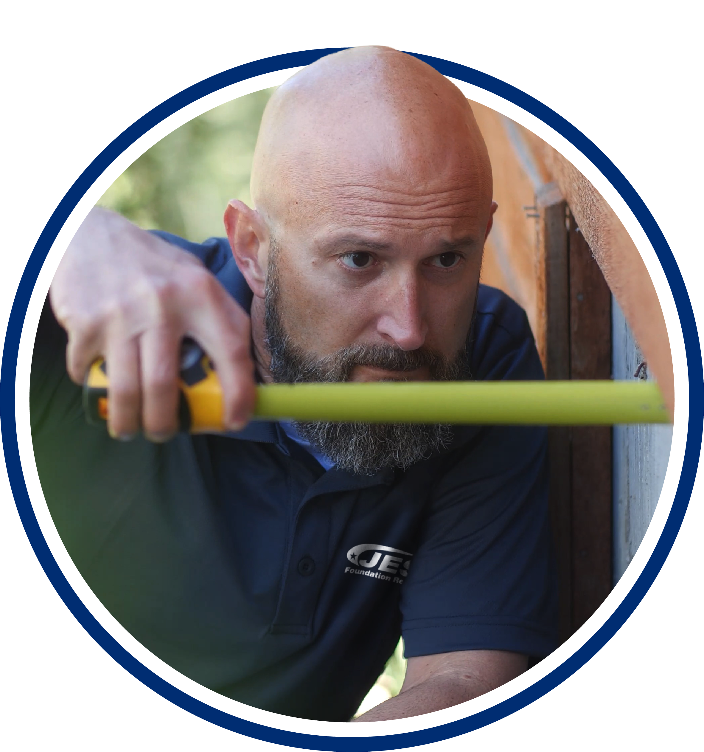 A man with a beard measures a wall with a tape measure, wearing a navy shirt with a company logo.