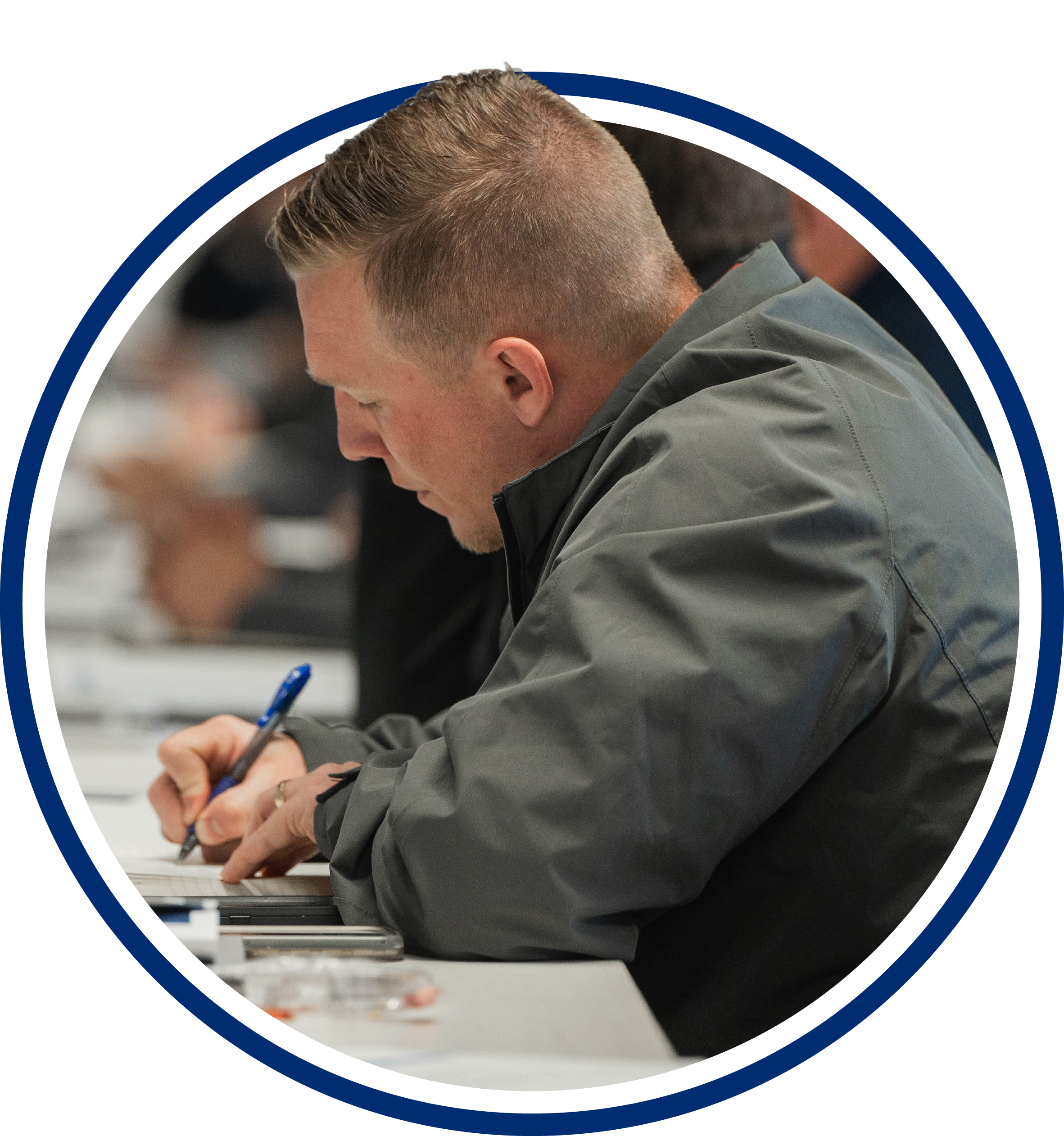 Man in a gray jacket writing with a pen at a desk, focused on his work in a classroom setting.