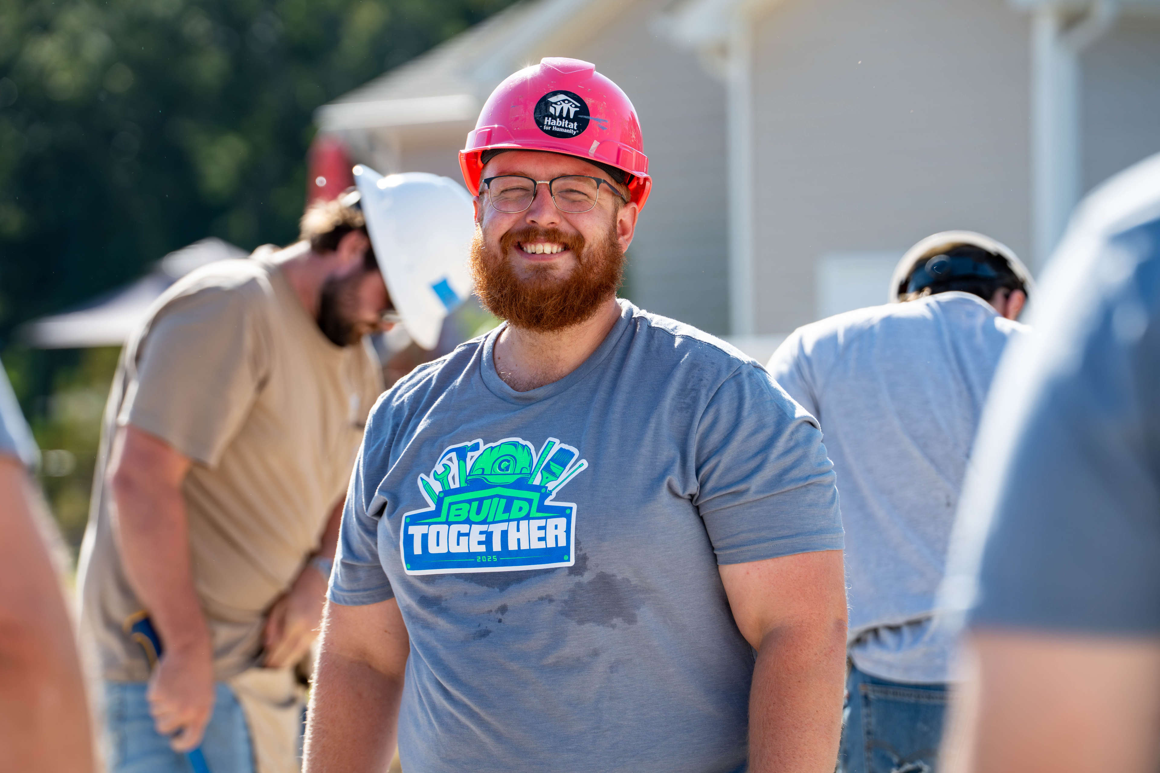 Smiling man in a red hard hat and "Build Together" shirt stands outdoors at a construction site with others working behind him.
