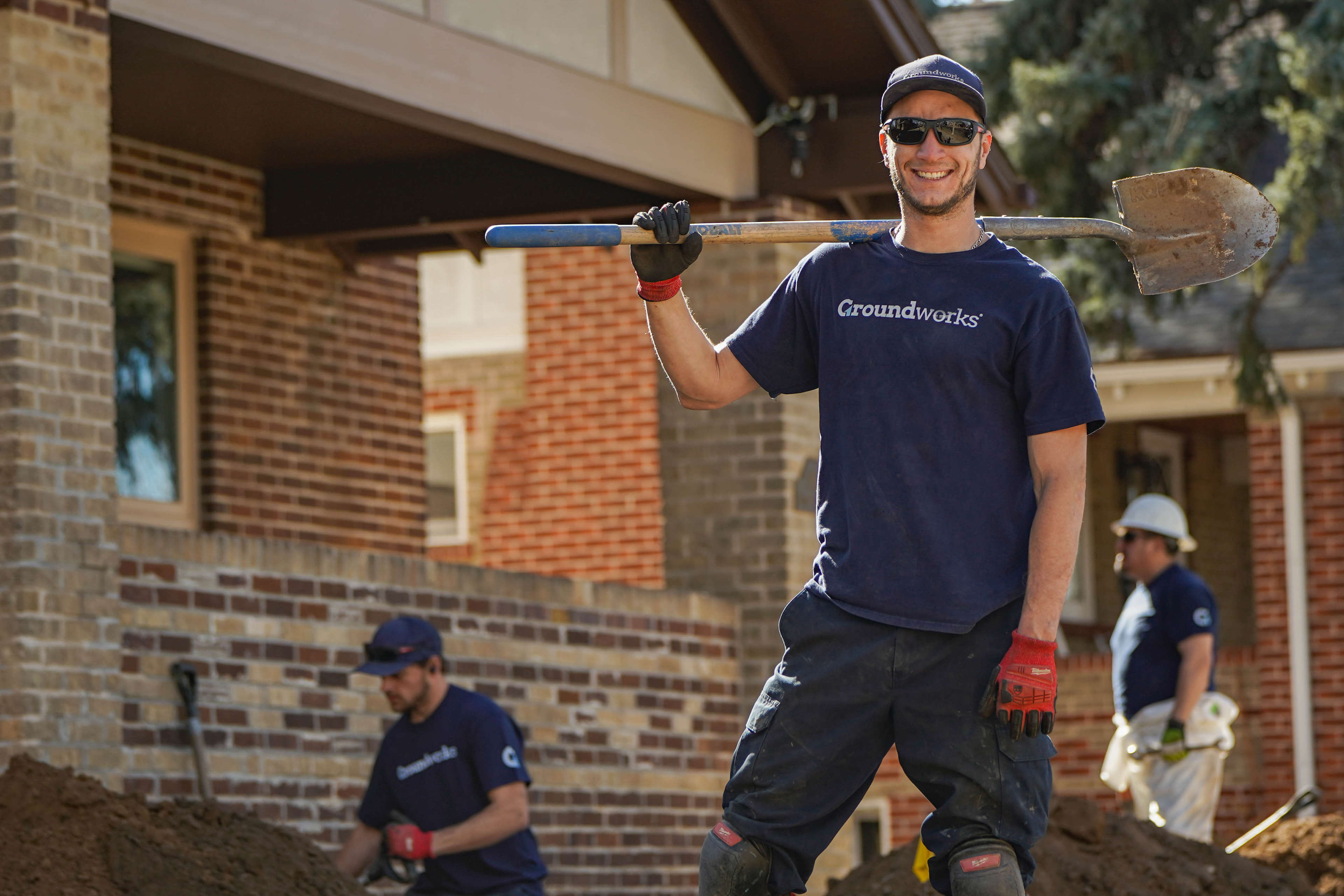 Smiling worker holding a shovel stands outside, with two coworkers digging near a brick house in the background.