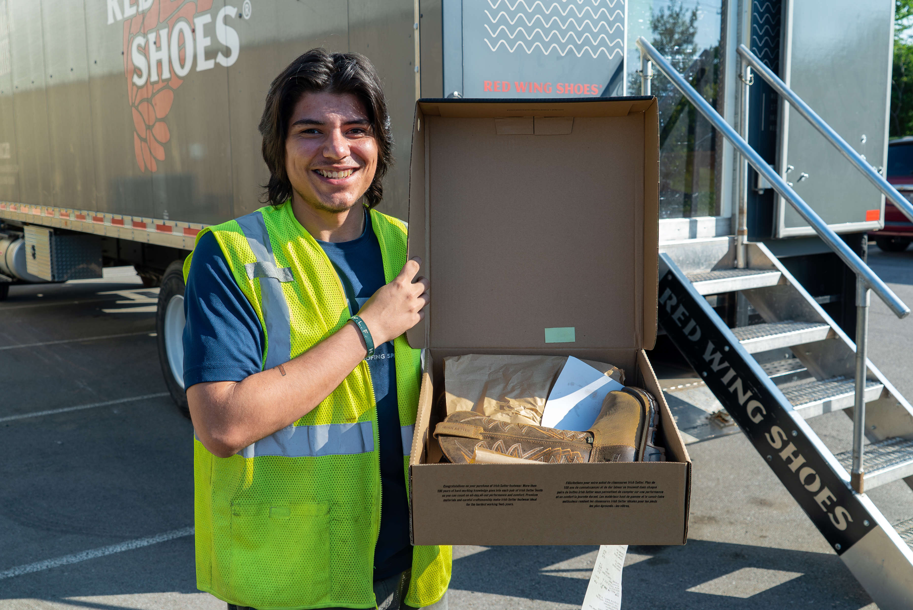 A smiling person in a safety vest holds an open shoebox with new boots in front of a "Red Wing Shoes" truck.
