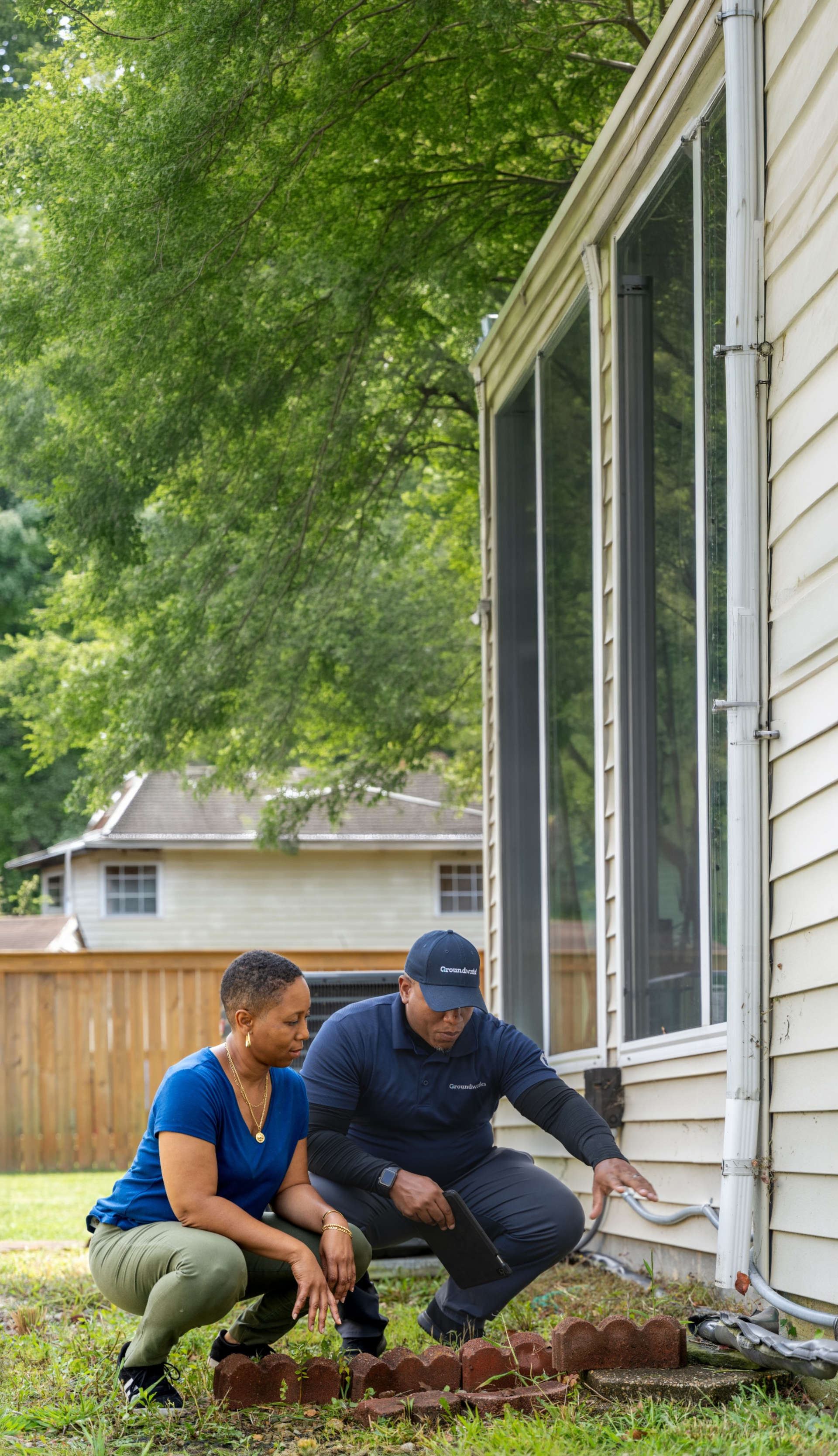 Two people crouch by a house inspecting bricks along the exterior wall, with trees and another house in the background.