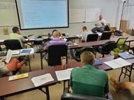 Classroom setting with people seated at desks, facing a whiteboard and large screen during a presentation.