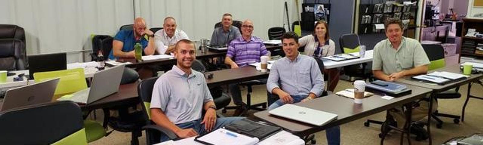 A group of people sitting at tables in a classroom setting, smiling at the camera.