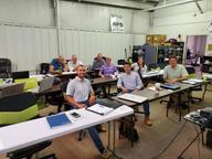 A group of people sitting at tables in a classroom setting, smiling at the camera.