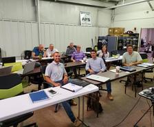 A group of people sitting at tables in a classroom setting, smiling at the camera.