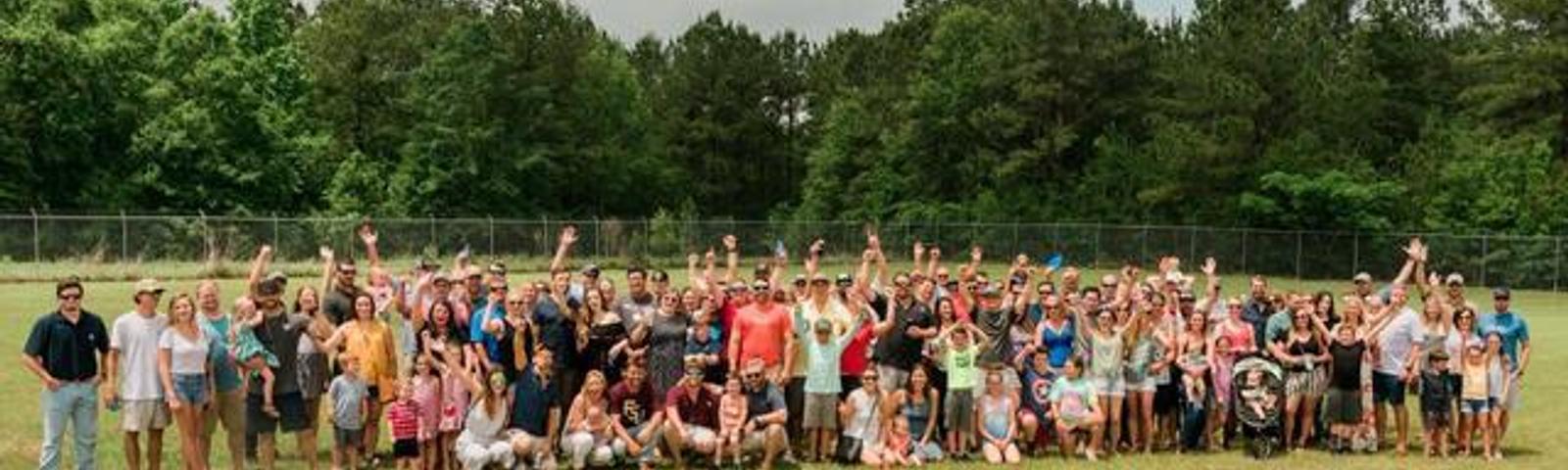 A large group of people posing outdoors with hands raised, surrounded by trees.