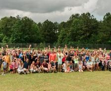 A large group of people posing outdoors with hands raised, surrounded by trees.