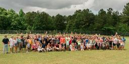A large group of people posing outdoors with hands raised, surrounded by trees.