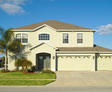 A two-story beige house with a three-car garage, palm tree, and clear blue sky.