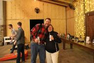 Two people smiling and holding drinks at a rustic indoor event with string lights and a cornhole game setup.