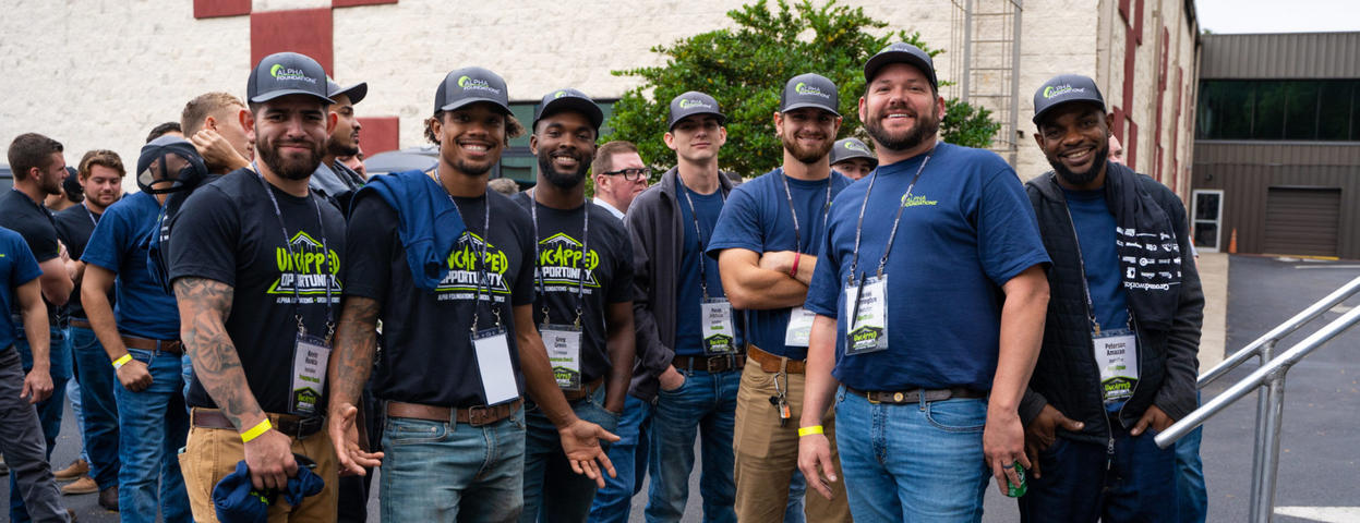 A group of smiling people in casual attire and hats pose outdoors near a building.