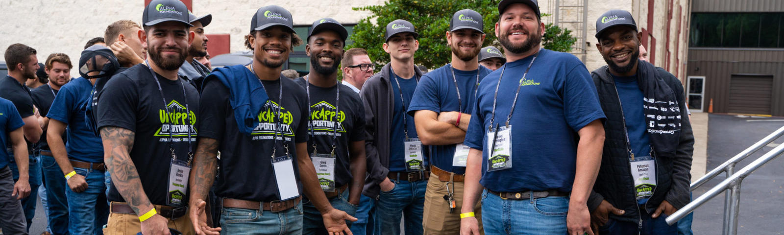 A group of smiling people in casual attire and hats pose outdoors near a building.