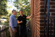 Two men inspecting a brick wall outside, one holding a tablet and pointing at the wall.