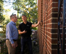 Two men inspecting a brick wall outside, one holding a tablet and pointing at the wall.