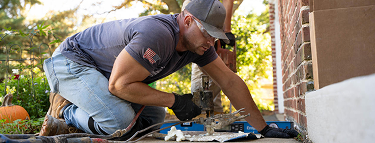 A man in a cap and gloves works with a tool outside, near a house.