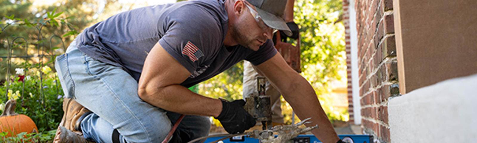 A man in a cap and gloves works with a tool outside, near a house.