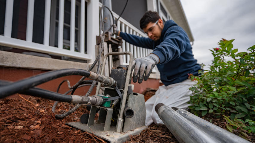 A person in gloves operates equipment near a house, surrounded by plants and soil.