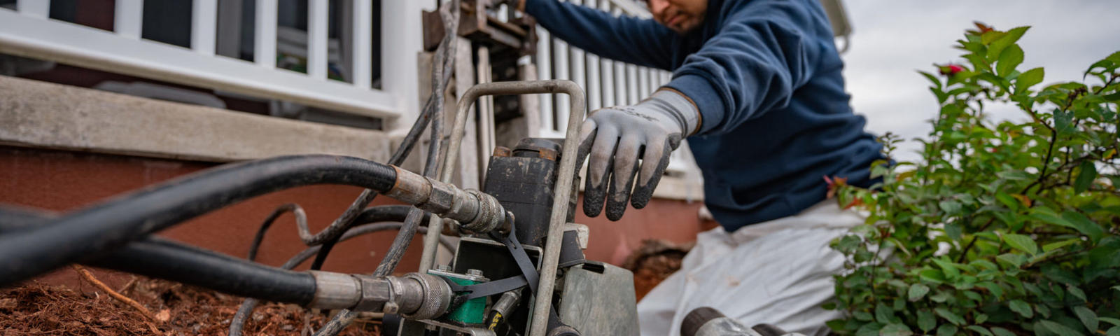 A person in gloves operates equipment near a house, surrounded by plants and soil.