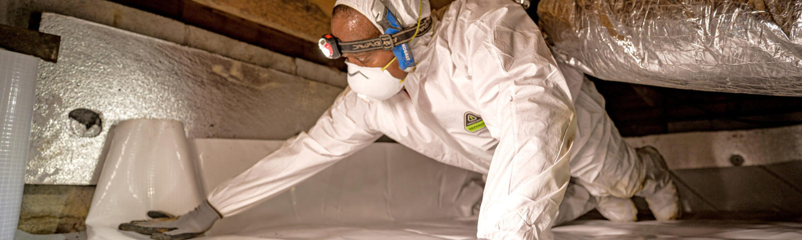 Person in protective gear installs insulation in a crawl space, wearing a headlamp and face mask.