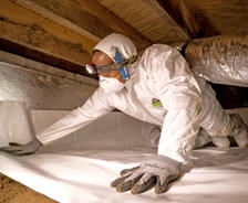 Person in protective gear installs insulation in a crawl space, wearing a headlamp and face mask.