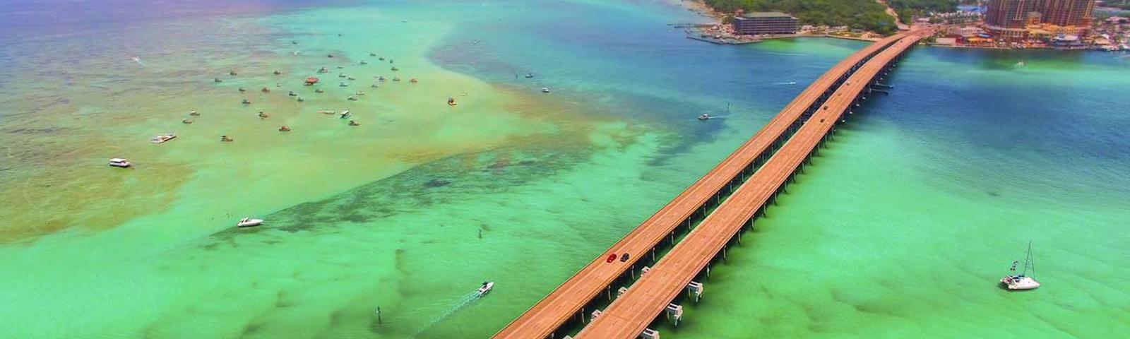 Aerial view of a long bridge over turquoise waters with several boats nearby and an island and buildings in the distance.