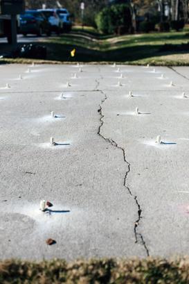 A cracked concrete sidewalk with pegs protruding stretches towards a grassy area, hinting at the need for concrete driveway repair, with parked cars visible in the background.