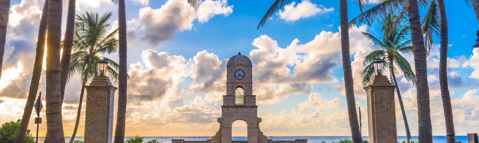 Palm trees frame a clock tower by the sea under a vibrant sky with scattered clouds.