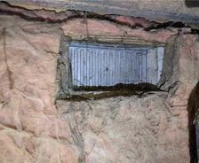 Rectangular vent embedded in a wall surrounded by pink insulation material and rough concrete surface.