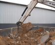 Excavator arm loading sand and debris into a large container next to a metal building.