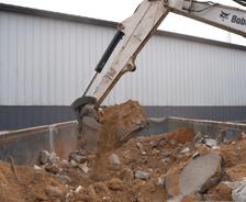 Excavator arm loading sand and debris into a large container next to a metal building.