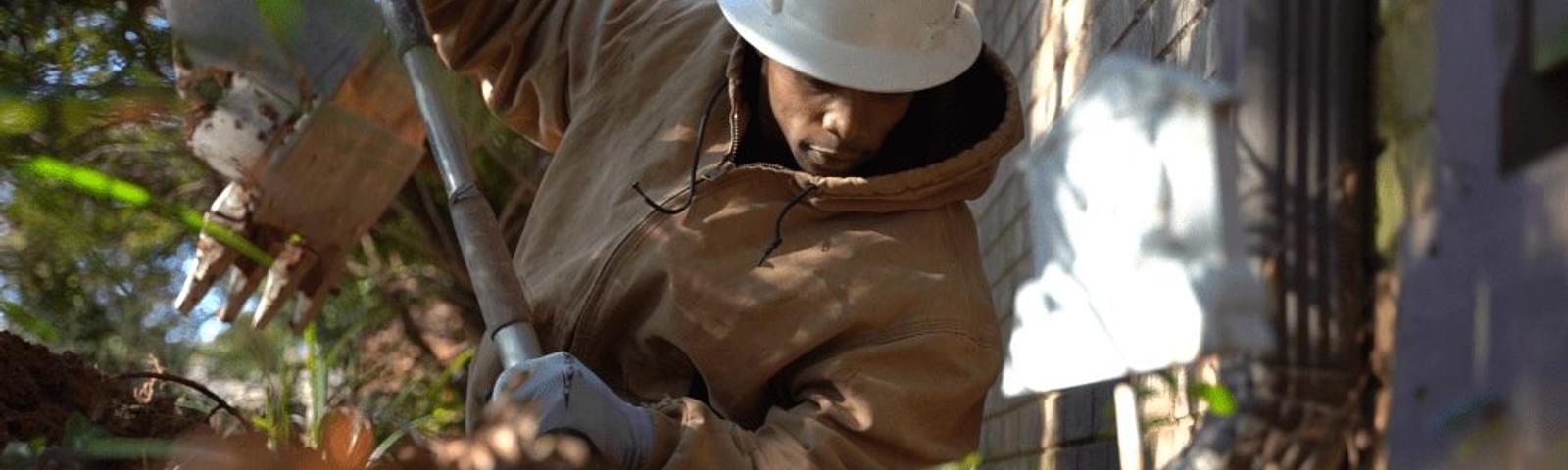 Person in a hard hat and jacket digging with a shovel near a building wall, surrounded by foliage.
