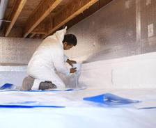 A person in protective gear applies insulation in a crawl space with wooden beams.